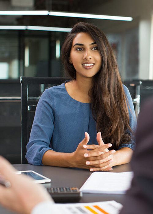 Portrait of young Indian female client or candidate sitting at table, talking to senior male manager and smiling in office. Job interview or consultancy concept